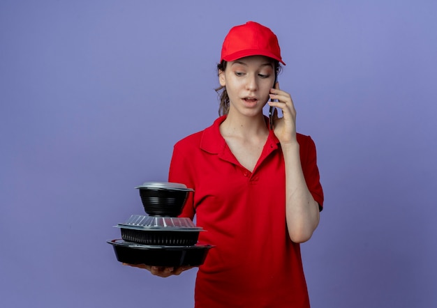 Impresionada joven bonita repartidora vestida con uniforme rojo y gorra sosteniendo y mirando contenedores de comida y hablando por teléfono aislado sobre fondo púrpura con espacio de copia