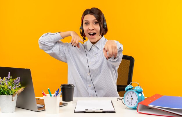 Impresionada joven call center chica con auriculares sentado en el escritorio apuntando y en su mano aislada en naranja