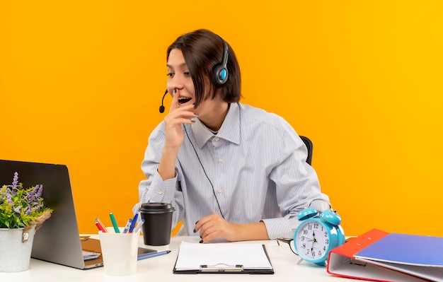 Impresionada joven call center chica con auriculares sentado en el escritorio mirando portátil con el dedo en los labios aislados en naranja