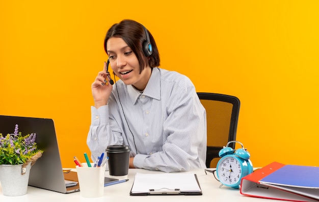 Impresionada joven call center chica con auriculares sentado en el escritorio poniendo la mano en el auricular aislado en naranja