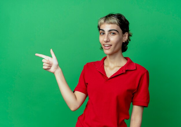 Impresionada joven caucásica con corte de pelo pixie apuntando al lado aislado sobre fondo verde