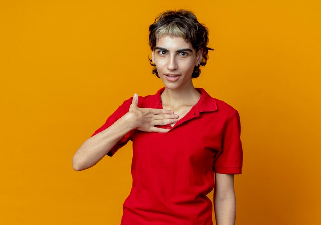 Impresionada joven caucásica con corte de pelo pixie poniendo la mano en el pecho aislado sobre fondo naranja con espacio de copia