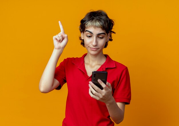 Impresionada joven caucásica con corte de pelo pixie sosteniendo y mirando el teléfono móvil con el dedo levantado