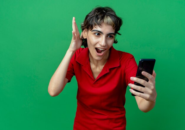 Impresionada joven caucásica con corte de pelo pixie sosteniendo y mirando el teléfono móvil y manteniendo la mano en el aire