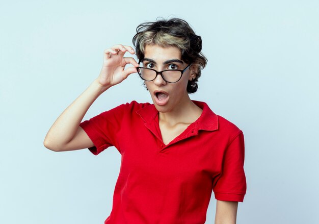 Impresionada joven caucásica con corte de pelo pixie vistiendo y sosteniendo gafas aisladas sobre fondo blanco.