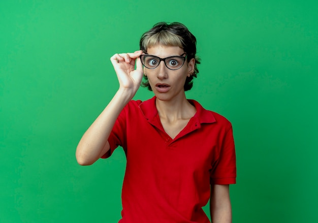 Impresionada joven caucásica con corte de pelo pixie vistiendo y sosteniendo gafas aisladas sobre fondo verde con espacio de copia