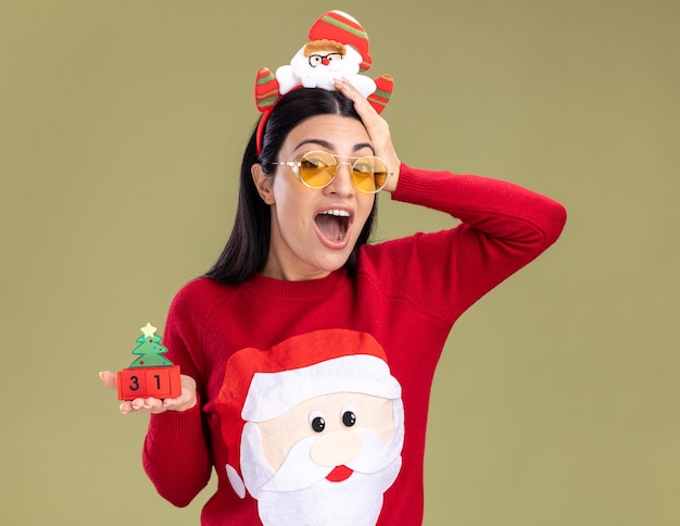 Impresionada joven caucásica con diadema de santa claus y suéter con gafas sosteniendo el juguete del árbol de Navidad con fecha mirando a cámara manteniendo la mano en la cabeza aislada sobre fondo verde oliva