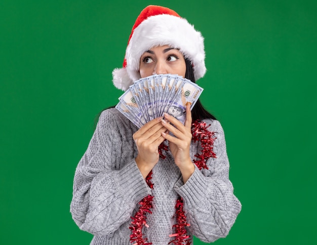 Impresionada joven caucásica con sombrero de navidad y guirnalda de oropel alrededor del cuello sosteniendo dinero mirando al lado desde atrás aislado en la pared verde con espacio de copia