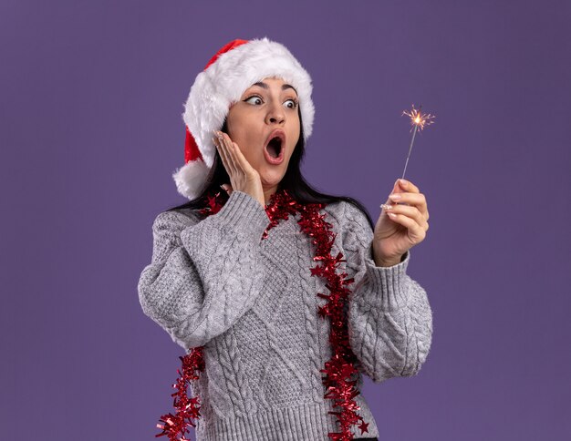 Impresionada joven caucásica con sombrero de navidad y guirnalda de oropel alrededor del cuello sosteniendo y mirando la bengala de vacaciones manteniendo la mano en la cara aislada sobre fondo púrpura