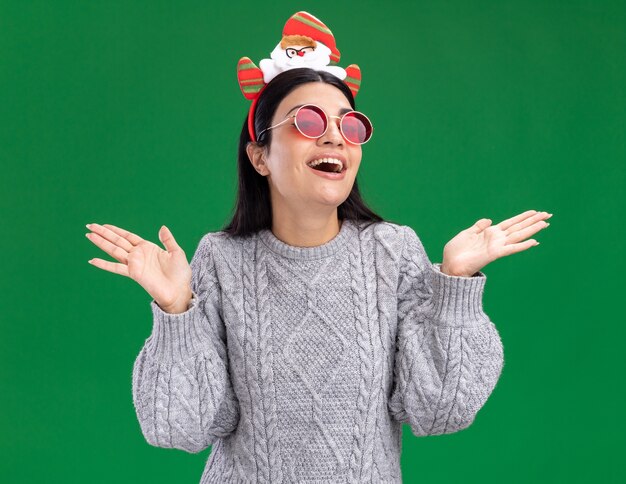 Impresionada joven caucásica vistiendo diadema de santa claus con gafas mostrando las manos vacías aisladas en la pared verde