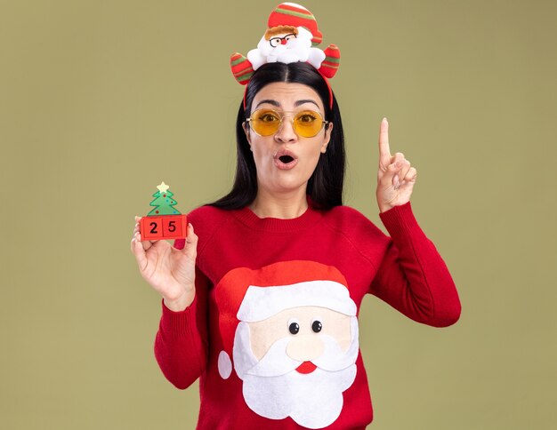 Impresionada joven caucásica vistiendo santa claus diadema y suéter con gafas sosteniendo el juguete del árbol de Navidad con fecha apuntando hacia arriba aislado en la pared verde oliva