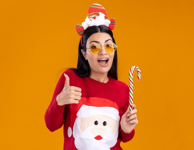 Impresionada joven caucásica vistiendo santa claus diadema y suéter con gafas sosteniendo tradicional bastón de caramelo navideño mostrando el pulgar hacia arriba aislado en la pared naranja con espacio de copia