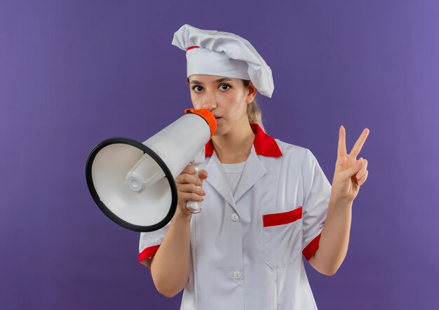Impresionada joven cocinera bonita en uniforme de chef hablando por altavoz y haciendo el signo de la paz aislado en la pared púrpura