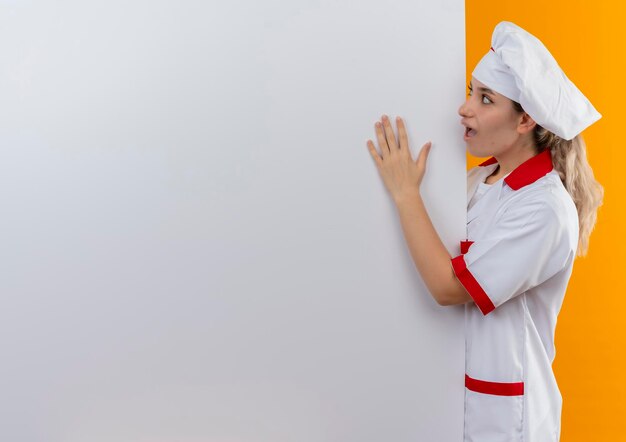 Impresionada joven cocinera bonita en uniforme de chef de pie detrás mirando y poniendo la mano en la pared blanca aislada en la pared naranja con espacio de copia