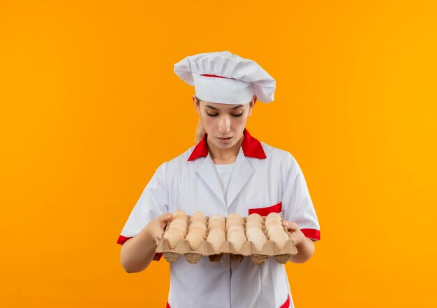 Impresionada joven cocinera bonita en uniforme de chef sosteniendo y mirando el cartón de huevos aislado en la pared naranja