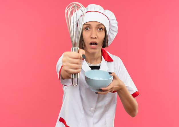 Impresionada joven cocinera en uniforme de chef estirando batidor y sosteniendo el tazón aislado en rosa con espacio de copia