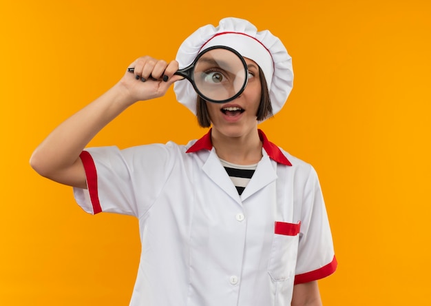 Impresionada joven cocinera en uniforme de chef mirando a través de lupa aislada en naranja