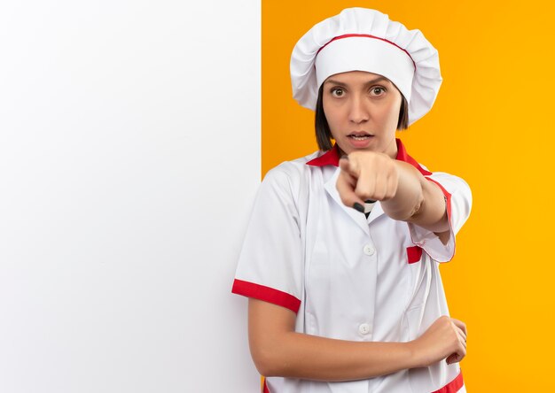 Impresionada joven cocinera en uniforme de chef de pie frente a una pared blanca y apuntando aislado en naranja con espacio de copia