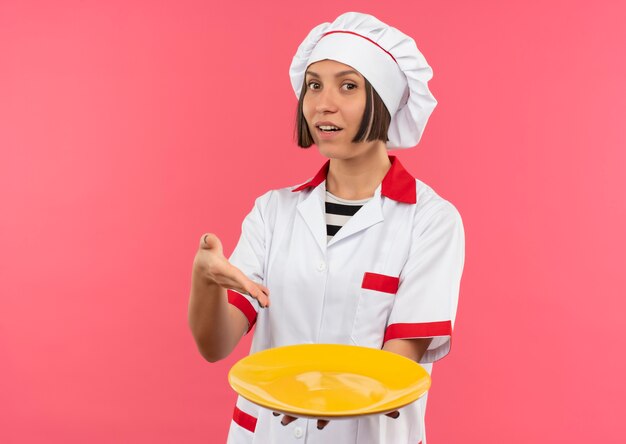 Impresionada joven cocinera en uniforme de chef sosteniendo y apuntando al plato vacío aislado en rosa con espacio de copia