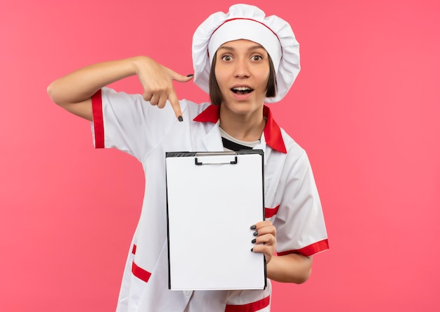 Foto gratuita impresionada joven cocinera en uniforme de chef sosteniendo y apuntando al portapapeles aislado en rosa con espacio de copia