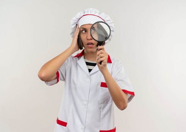 Foto gratuita impresionada joven cocinera en uniforme de chef sosteniendo lupa poniendo la mano en la cabeza y mirando al lado aislado en blanco con espacio de copia