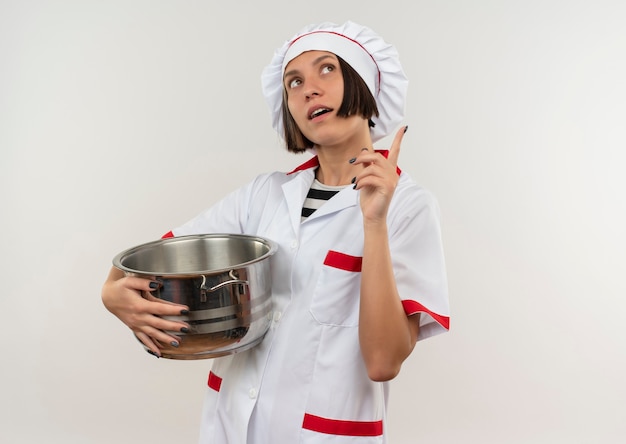 Impresionada joven cocinera en uniforme de chef sosteniendo la olla levantando el dedo y mirando hacia arriba aislado en blanco con espacio de copia