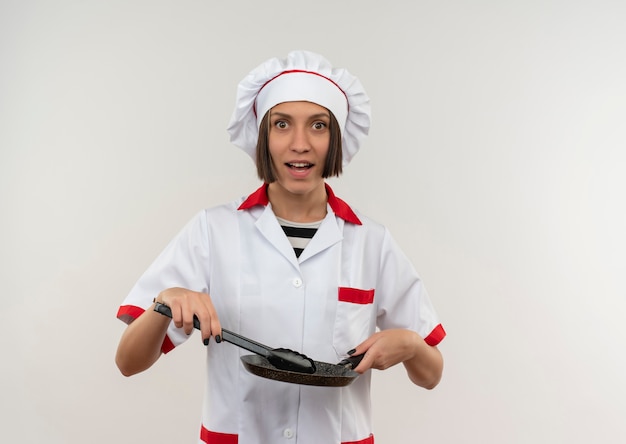 Impresionada joven cocinera en uniforme de chef sosteniendo pinzas y sartén aislado en blanco con espacio de copia