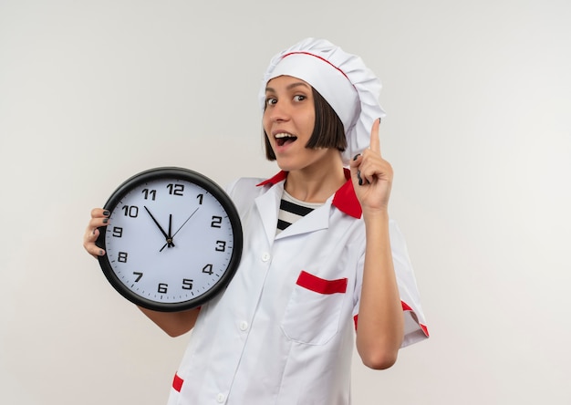 Impresionada joven cocinera en uniforme de chef sosteniendo el reloj y levantando el dedo aislado en blanco con espacio de copia