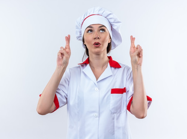 Impresionada joven cocinera vistiendo uniforme de chef cruzando los dedos aislado en la pared blanca