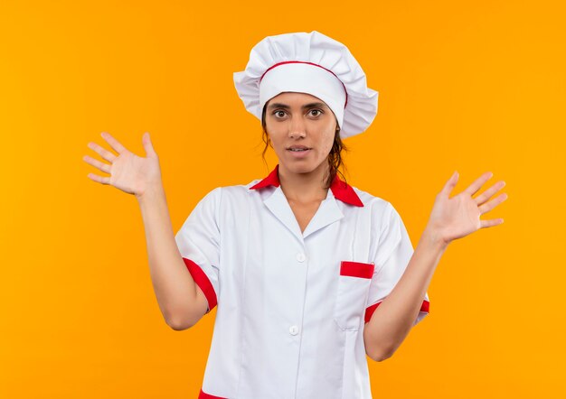Impresionada joven cocinera vistiendo uniforme de chef extiende las manos sobre la pared amarilla aislada con espacio de copia