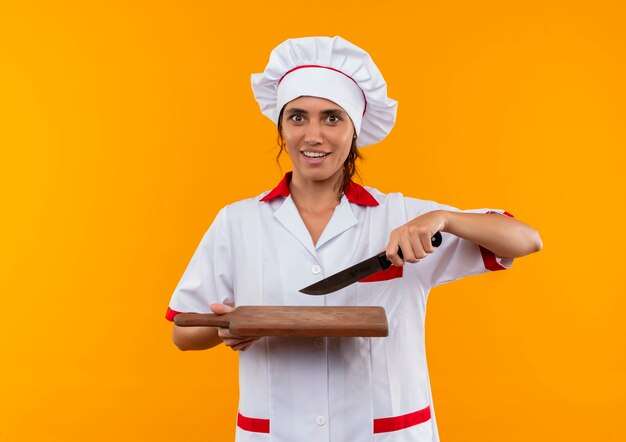 Impresionada joven cocinera vistiendo uniforme de chef sosteniendo un cuchillo y una tabla de cortar en la pared amarilla aislada con espacio de copia