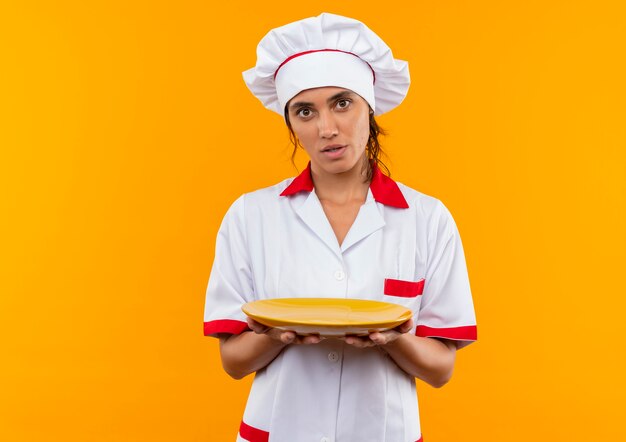 Impresionada joven cocinera vistiendo uniforme de chef sosteniendo la placa en la pared amarilla aislada con espacio de copia