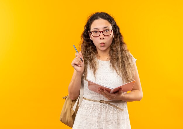 Impresionada joven colegiala bonita con gafas y bolsa trasera con bolígrafo y bloc de notas aislado en amarillo