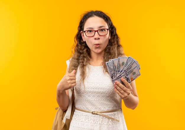 Impresionada joven colegiala bonita con gafas y bolsa trasera con dinero aislado en amarillo