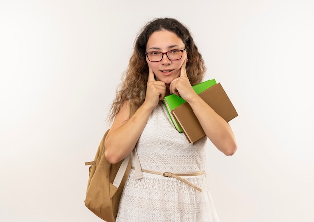 Impresionada joven colegiala bonita con gafas y bolsa trasera sosteniendo libros mirando al frente poniendo los dedos en la cara aislada en blanco