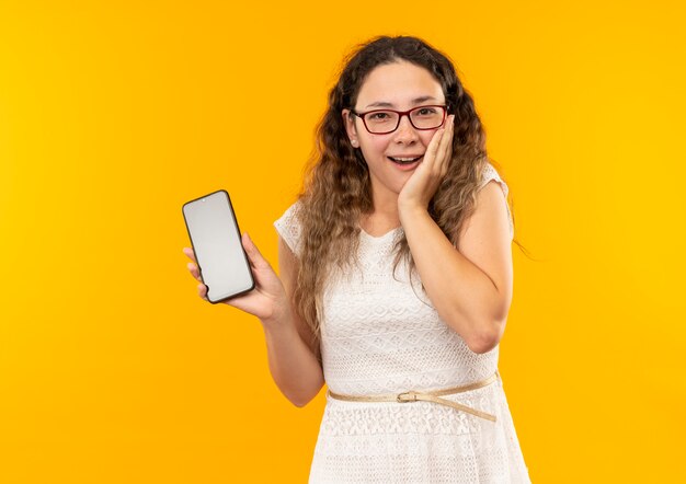 Impresionada joven colegiala bonita con gafas y bolso trasero sosteniendo el teléfono móvil poniendo la mano en la mejilla aislada en amarillo