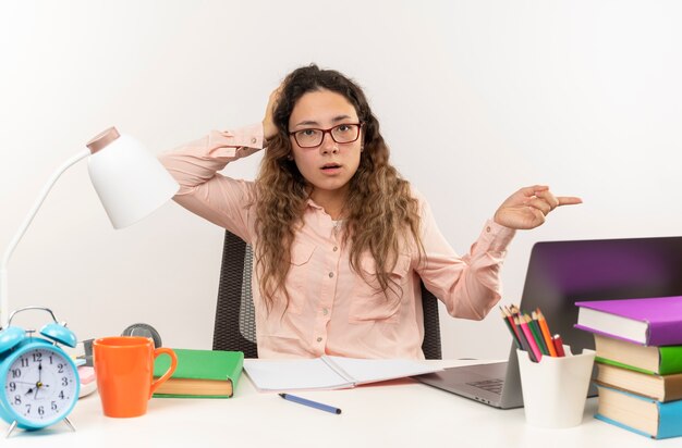 Impresionada joven colegiala bonita con gafas sentado en el escritorio con herramientas escolares haciendo sus deberes poniendo la mano en la cabeza y apuntando al lado aislado en blanco