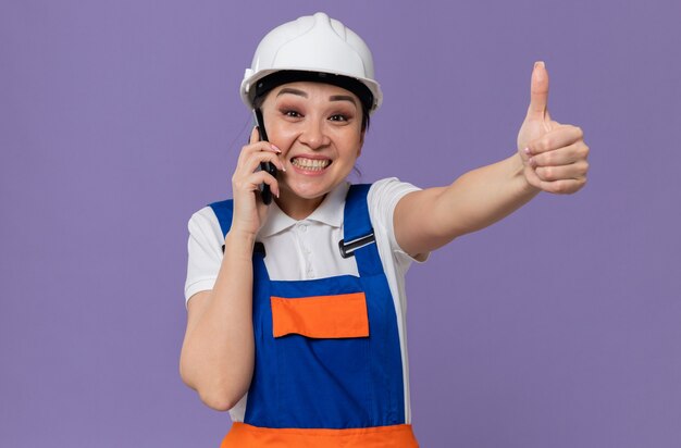 Impresionada joven constructora asiática con casco de seguridad blanco hablando por teléfono y apuntando hacia arriba