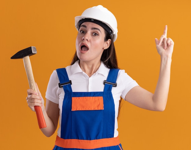 Impresionada joven constructora en uniforme sosteniendo puntos de martillo en arriba aislado en la pared naranja