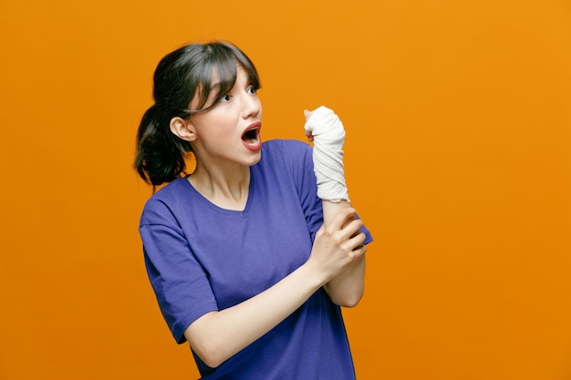 Impresionada joven deportista con camiseta girando la cabeza hacia un lado mirando a un lado agarrando su brazo con la muñeca envuelta con una venda aislada en un fondo naranja