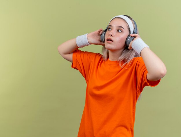 Impresionada joven deportista caucásica con tirantes en los auriculares con diadema y muñequeras mira al lado