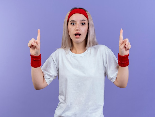 Impresionada joven deportiva con tirantes con diadema y muñequeras apunta hacia arriba con las dos manos aisladas en la pared púrpura