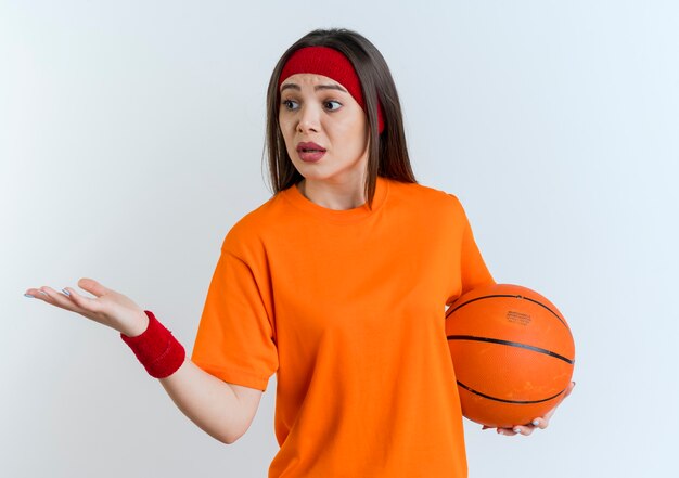 Impresionada joven deportiva vistiendo diadema y muñequeras mostrando la mano vacía mirando al lado sosteniendo la pelota de baloncesto