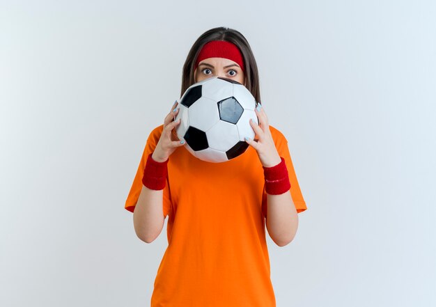 Foto gratuita impresionada joven deportiva vistiendo diadema y muñequeras sosteniendo un balón de fútbol mirando desde atrás aislado