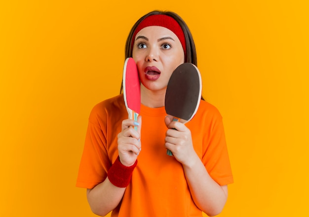 Impresionada joven deportiva vistiendo diadema y muñequeras sosteniendo raquetas de ping pong tocando la cara con ellos mirando al lado