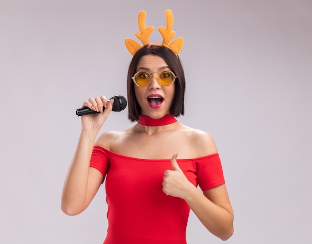 Impresionada joven con diadema de cuernos de reno y gafas sosteniendo el micrófono mirando a la cámara mostrando el pulgar hacia arriba aislado sobre fondo blanco.