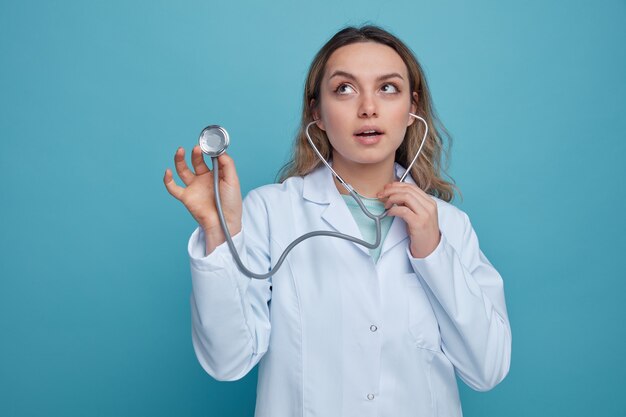 Impresionada joven doctora vistiendo bata médica y estetoscopio agarrando estetoscopio mirando hacia arriba