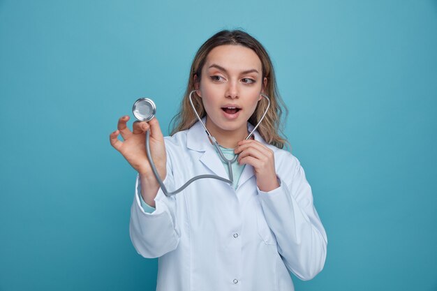 Impresionada joven doctora vistiendo bata médica y un estetoscopio agarrando y mirando el estetoscopio