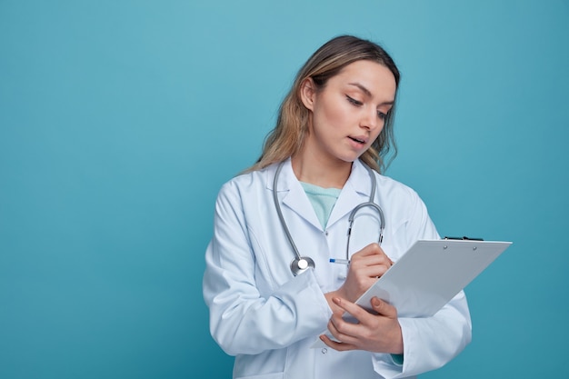 Impresionada joven doctora vistiendo bata médica y estetoscopio alrededor del cuello escribiendo con lápiz en el portapapeles