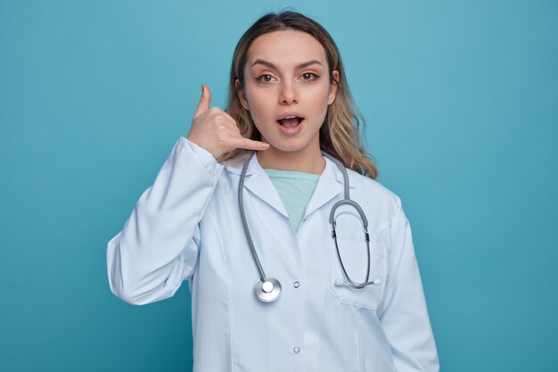 Impresionada joven doctora vistiendo bata médica y estetoscopio alrededor del cuello haciendo gesto de llamada